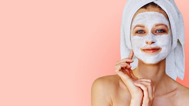 Pink background with a woman with a towel around her hair and index finger touching her skincare face mask. Preventing acne/breakouts on oily skin. Habits you should and shouldn't do. Clean skincare 