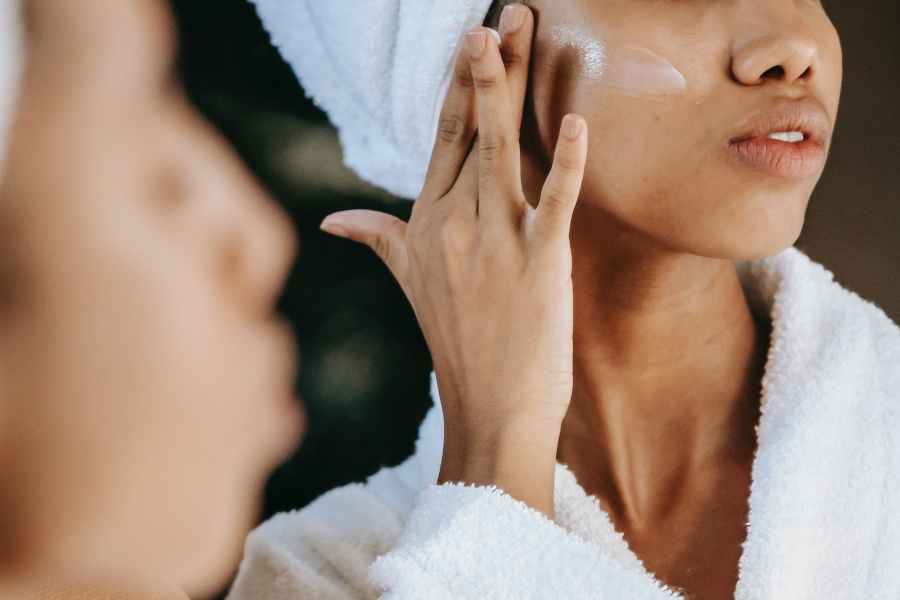 Woman in white robe and towel wrap on hair putting a finger of skincare product on face.